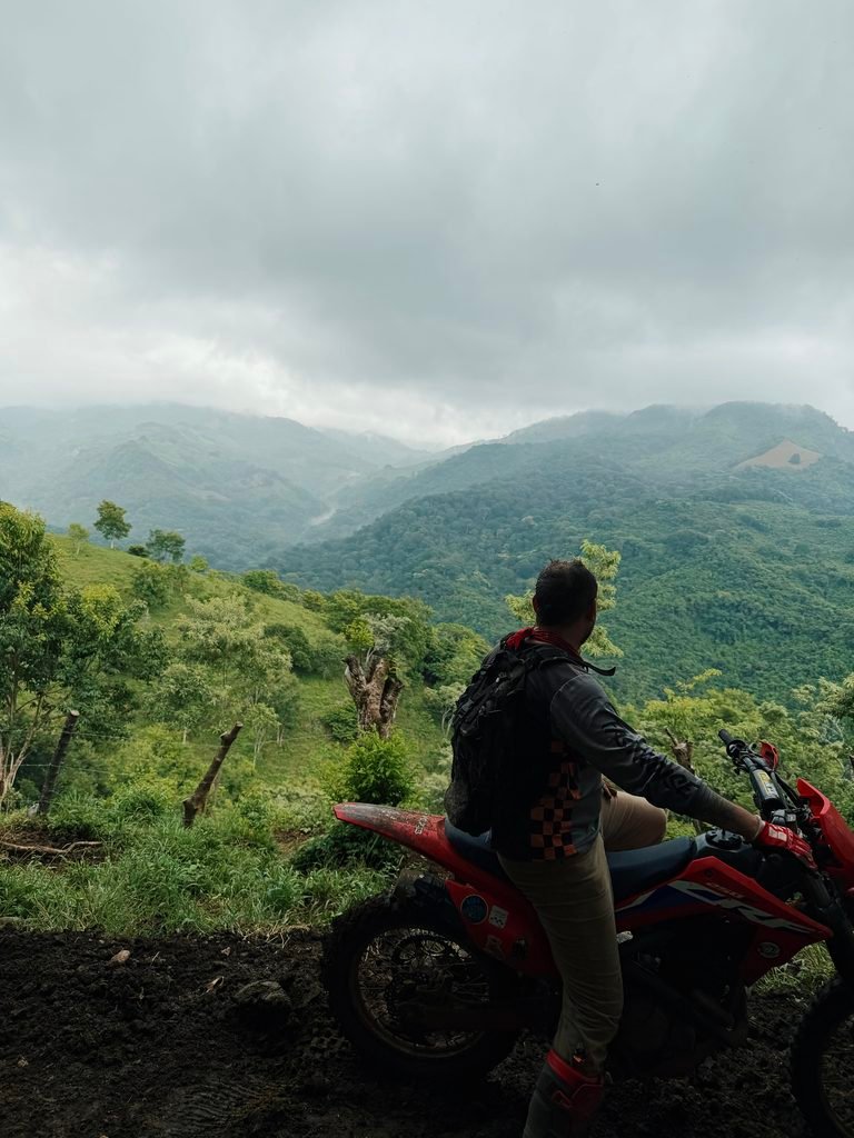 Rider on a red dirt bike looking out over green mountains under cloudy skies in El Salvador