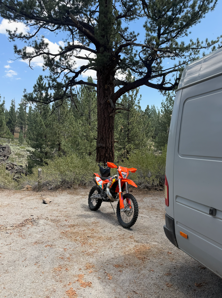 KTM dirt bike parked next to the Sprinter moto van under a pine tree at a campsite near Mammoth